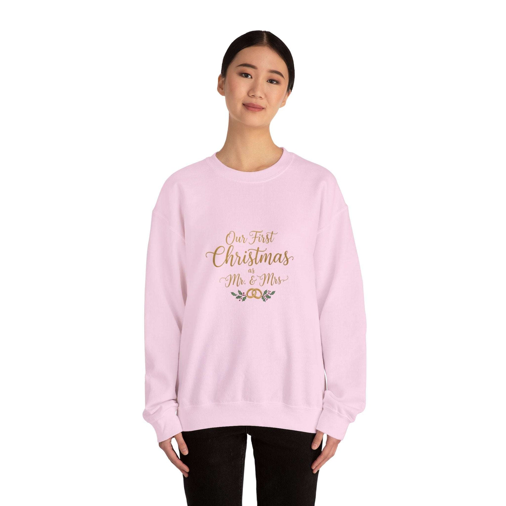 His/Hers Newlyweds Sweatshirt in light pink with 'Our First Christmas Mr. & Mrs.' gold embroidery. Cozy unisex crewneck ideal for holiday gifting.